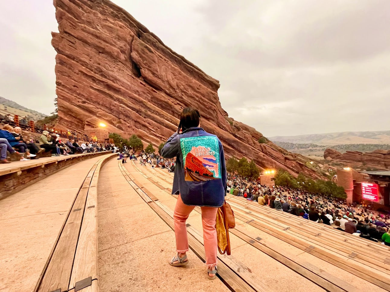 photo of a woman at red rocks stadium wearing a custom chainstitched denim jacket stitched with a sunset, red rocks, and text that says "I Have Dreams"