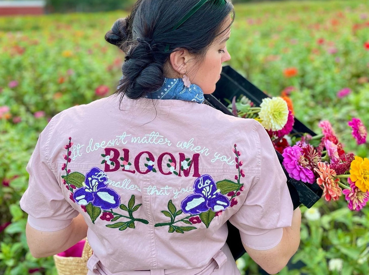 photo of a woman in a flower field wearing a custom chainstitched lavendar jumpsuit, stitched with wildflowers and song lyrics of "it doesn't matter when you bloom, it matters that you do".  