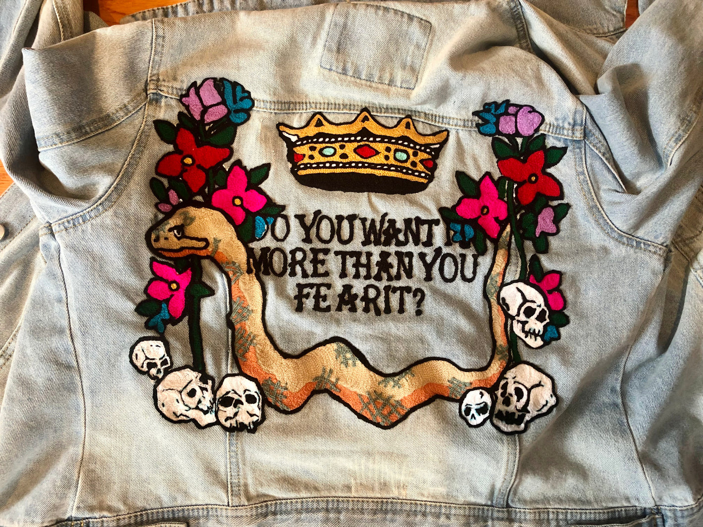 photo of a chainstitched denim jacket with a snake, skulls, flowers, and crown and text that says "do you want it more than you fear it?"